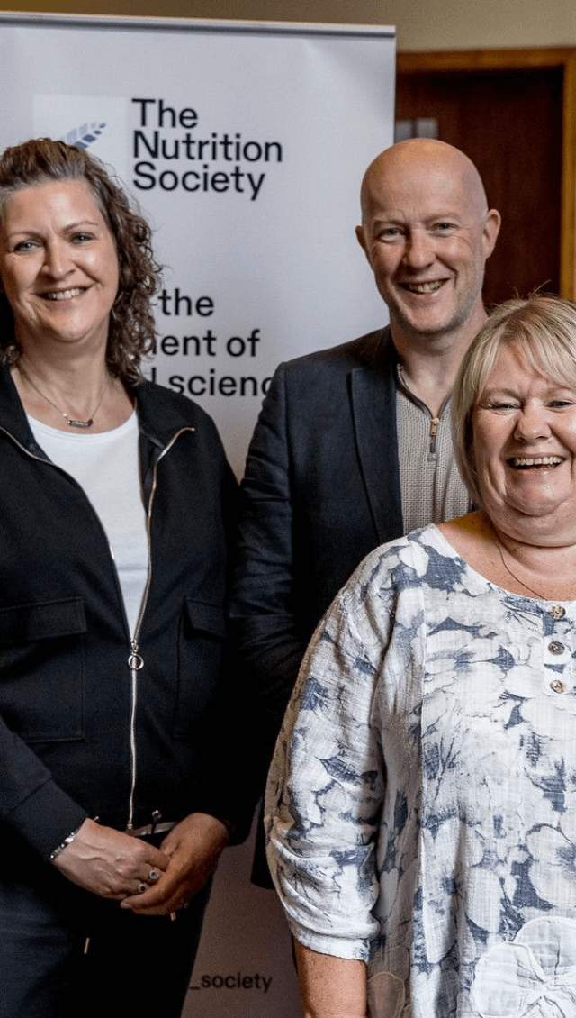 People standing smiling at the camera, in front of a pop up banner that reads 'The Nutrition Society'