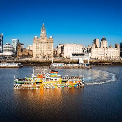 A sunny day in Liverpool. The image shows the Mersey Ferry which has been painted in bright colours and patterns on the River Mersey in front of the Royal Liver Building.