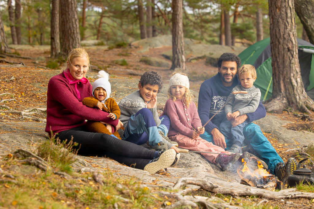 En familie på seks som sitter rundt bålet og griller pølser. Foto.
