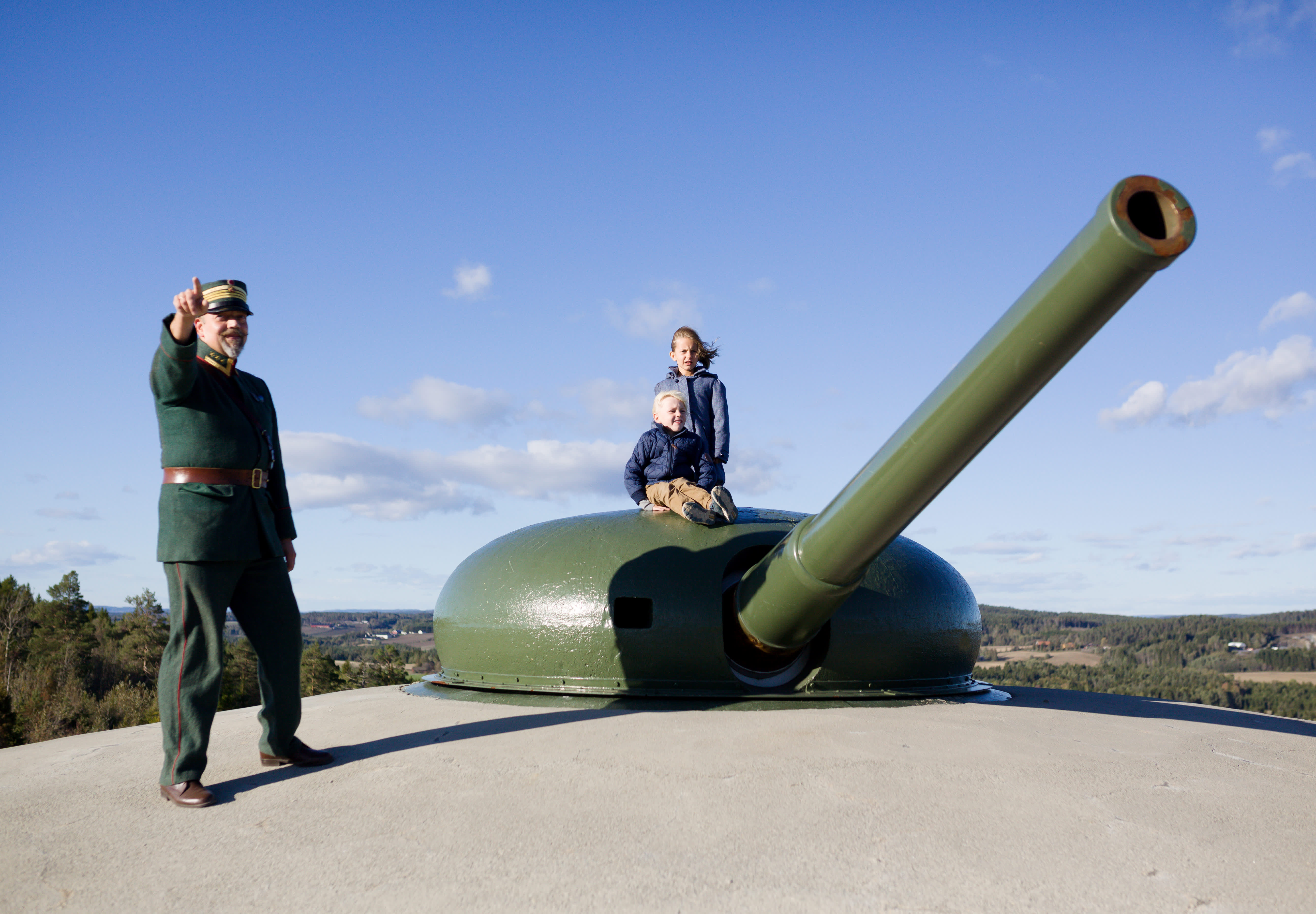 En uniformert mann peker utover ved kanonen på Høytorp fort, to barn sitter på kanonen