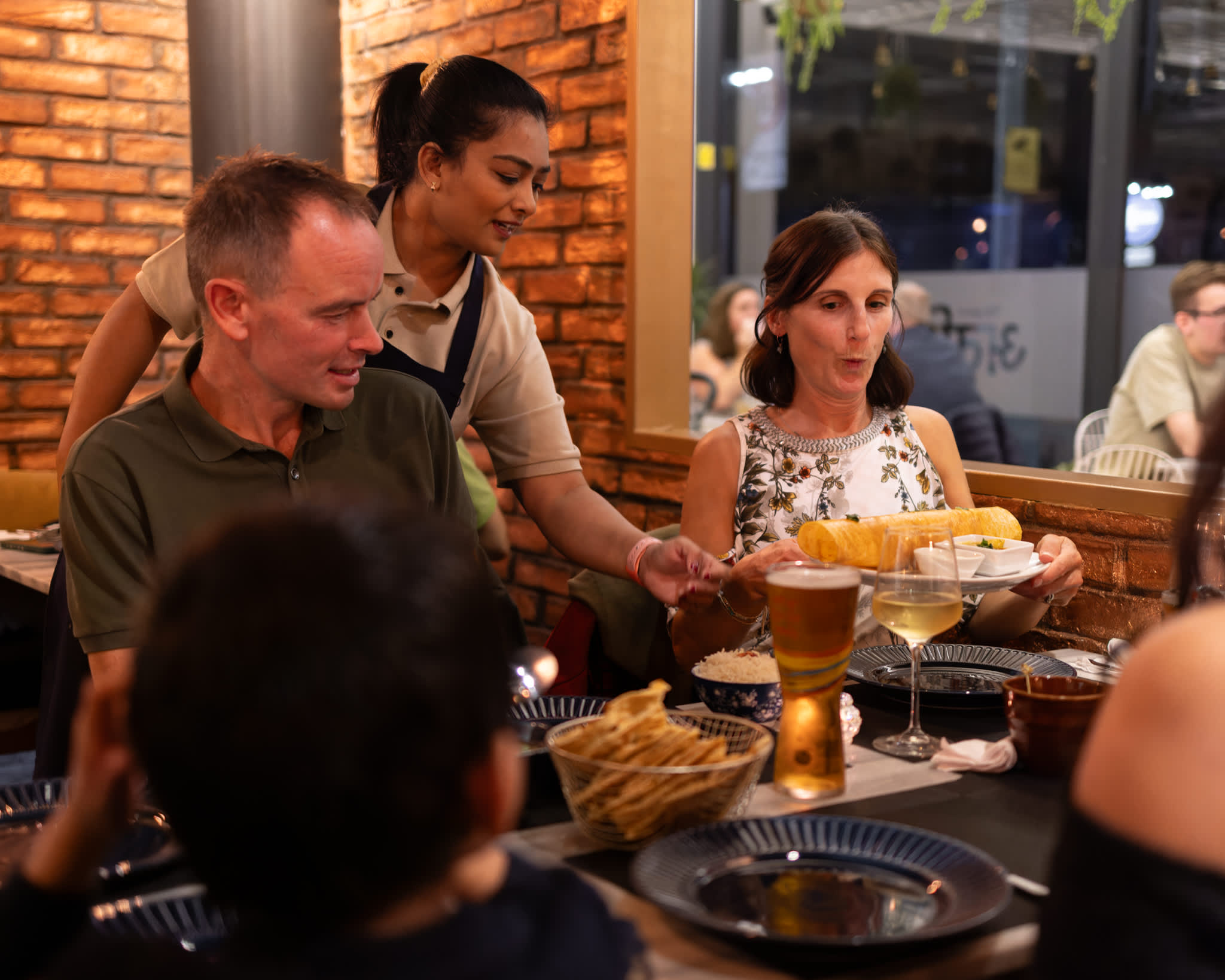 Waitress serving vegetarian Indian dish to guests seated at table as woman smiles seeing the dish