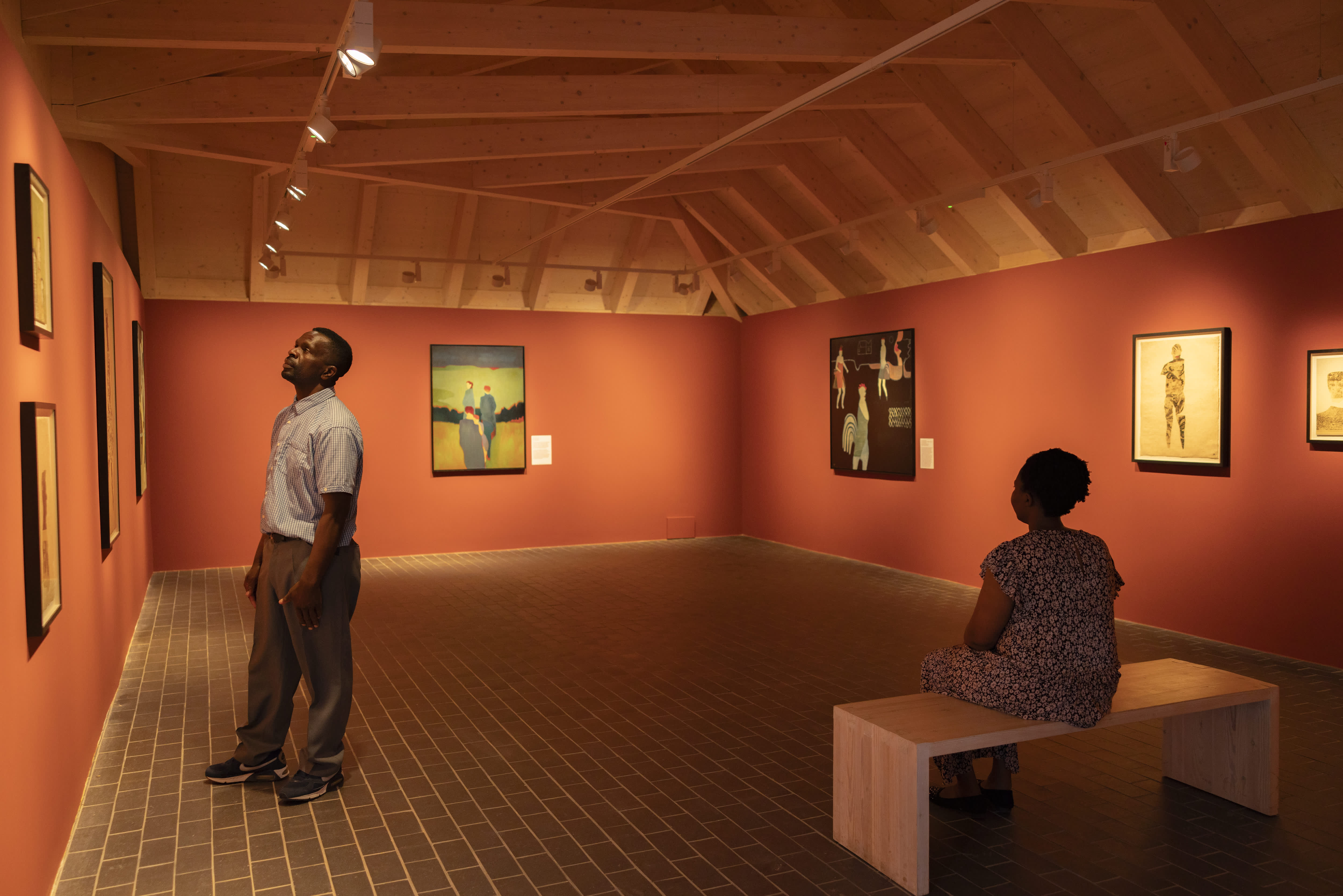 A couple stand in the gallery at Charleston, looking at artwork hanging on orange colour walls
