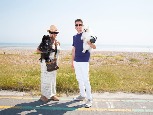 Two people standing in the sunshine alongside East Beach holding their cute, fluffy  dogs