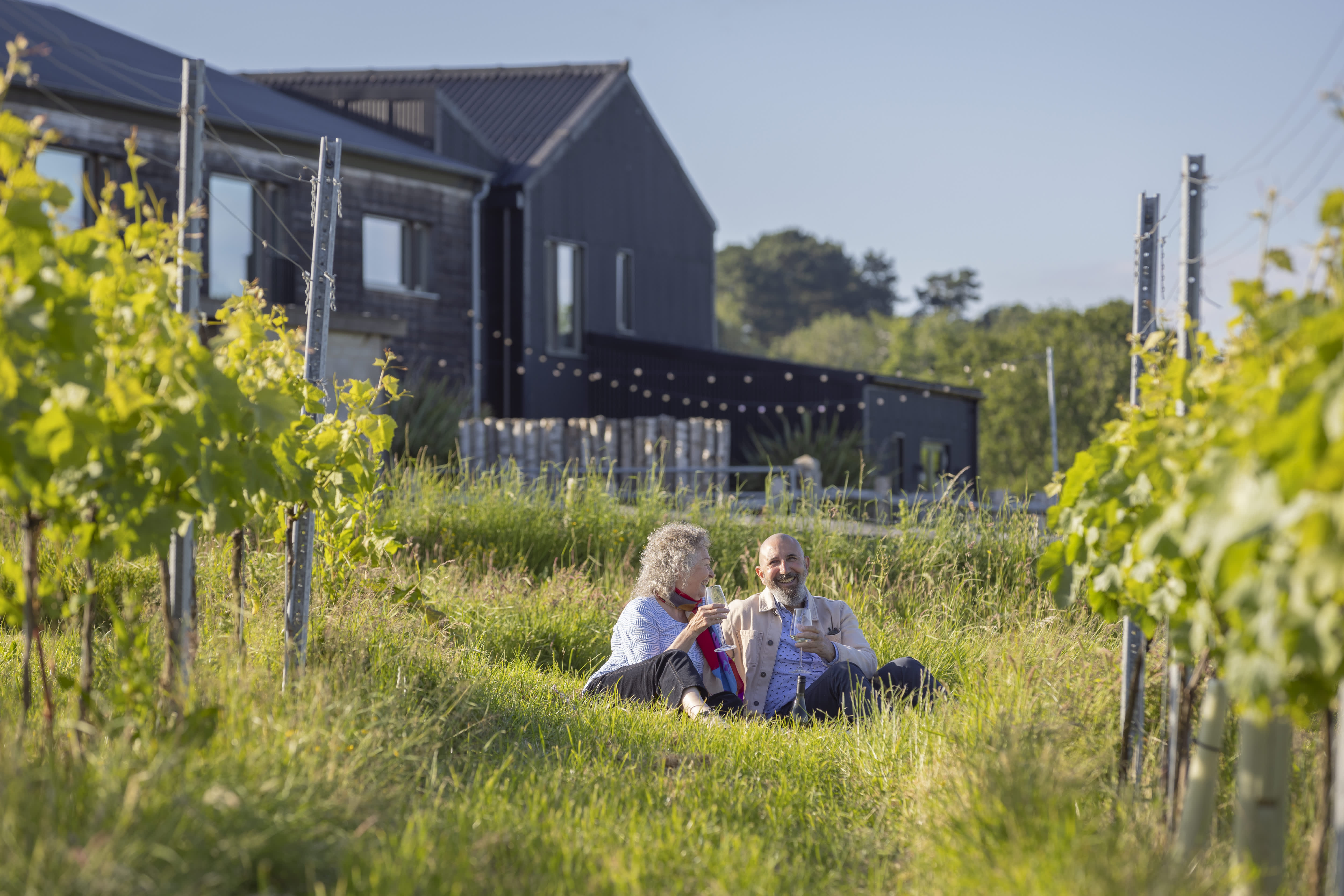 A couple sit in the vines with a glass of wine at Tillingham vineyard, with black barn buildings behind