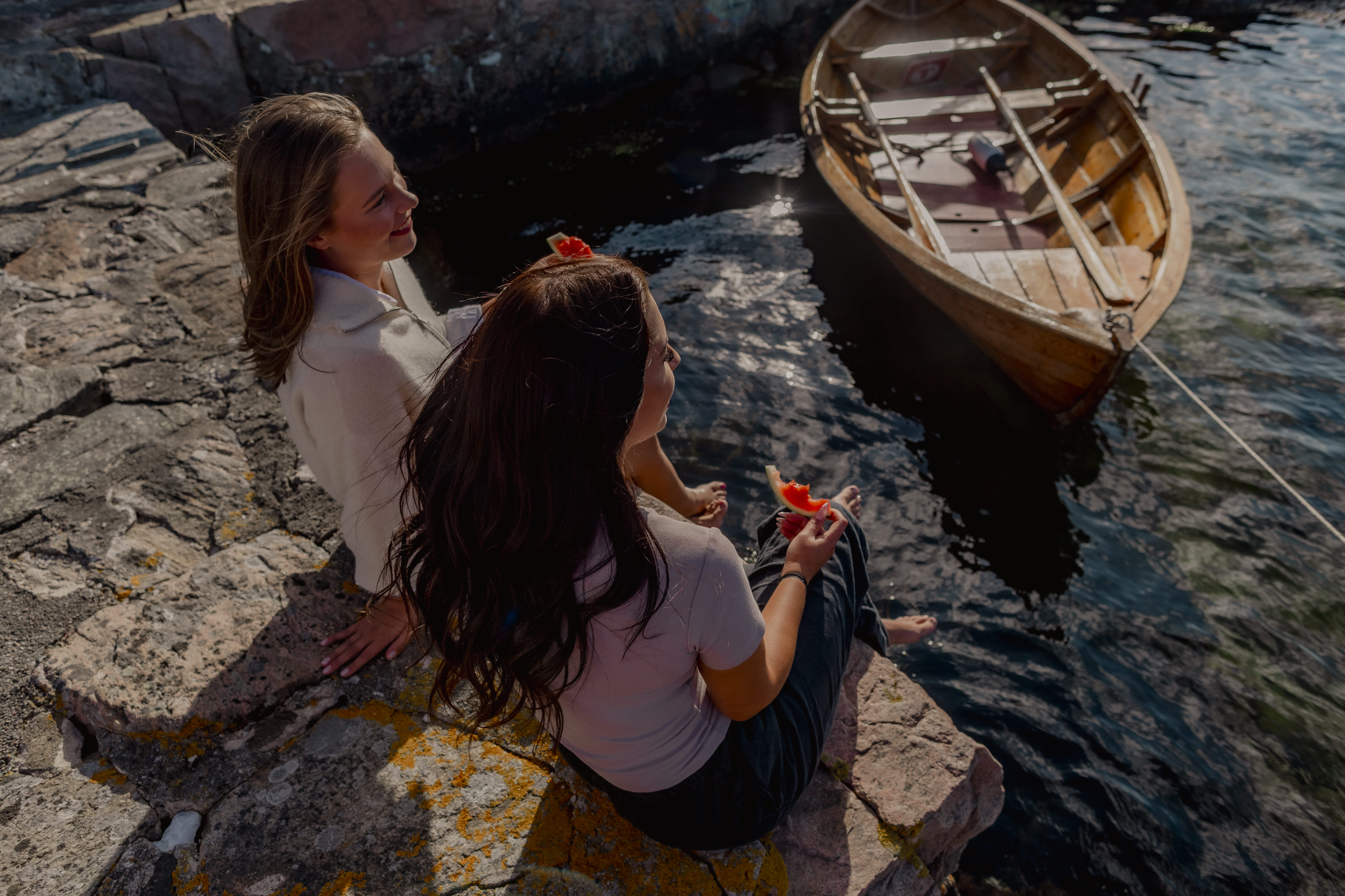 To kvinner spiser vannmelon på en brygge. Robåt i tre og havet i bakgrunnen