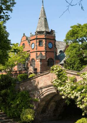 A red brick bridge covered in greenery. Behind is a large red brick church with a turret roof and clock face.