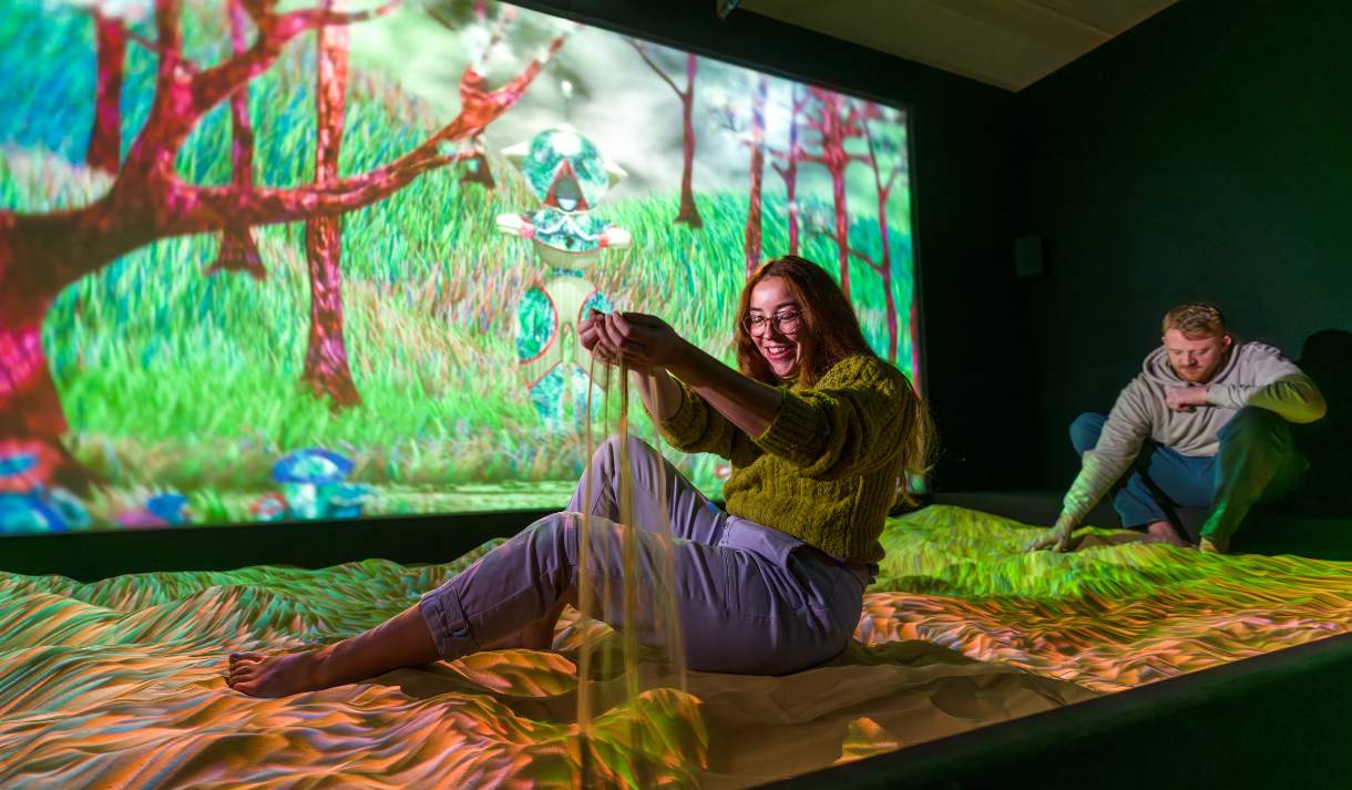 A lady sits in a sandpit in a dark room with a large, projected screen of nature behind her