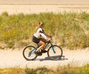 A girl rides a moutain bike on a trail path next to sand dunes and the beach on a sunny day