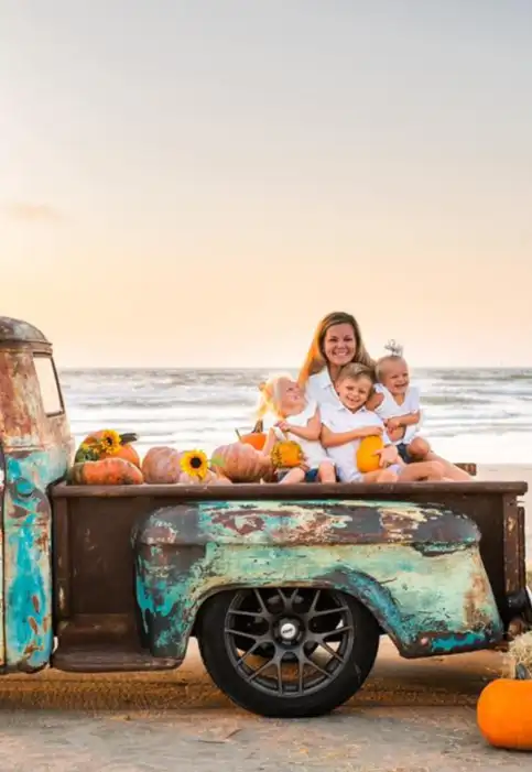 An old truck sits on the beach surrounded by pumpkins. A family sits in the bed of the truck and a logo overlaid on the photo reads "Beachtober."