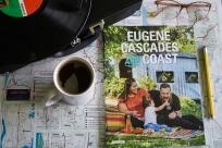 A visitor guide showing a family picnicking near a covered bridge sits on a table with a map, cup of coffee, glasses, a pencil and a record player.