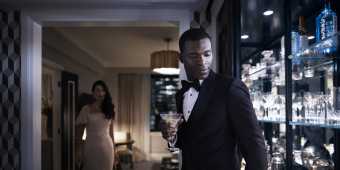 A man in a suit holding a wine glass with a woman in a long dress walks to him at the Fairmont Royal York hotel room in Toronto