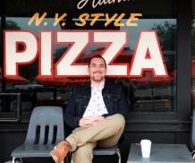 Man sitting on plastic chair in front of painted window with the words "N.Y. Style Pizza".