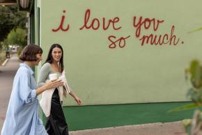 Two women walking in front of the I Love You So Much mural with coffee in hand.
