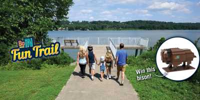 Family walking down a riverside path toward an overlook on the Ohio River as part of the SoIN Fun Trail, with a graphic promoting a buildable bison prize.
