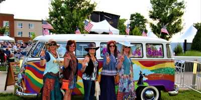 five women in hippie clothing giving peace sign stand in front of a brightly painted VW bus