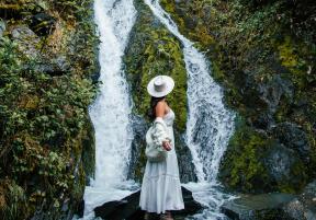 a woman in a brimmed hat and dress standing in front of a waterfall