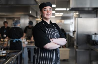 A chef standing with their arms folded and smiling in the centre of a commercial kitchen.