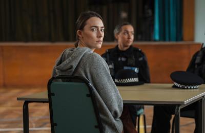 A woman in a grey hoodie sits at a table in a large hall, looking towards the camera. Two uniformed police officers sit across the table, with two police hats placed in front of them.