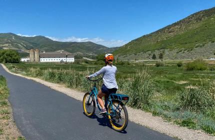 Woman riding an electric bike along a paved path with a white barn in the background