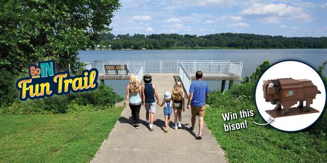 Family walking down a riverside path toward an overlook on the Ohio River as part of the SoIN Fun Trail, with a graphic promoting a buildable bison prize.