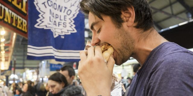 A man eating a peameal bacon sandwich with a Toronto Maple Leafs flag behind him