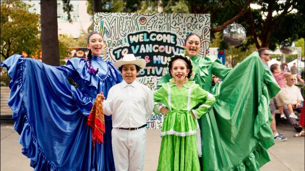Performers wearing elaborate blue, white and green clothes in front of a sign stating "Welcome to Vancouver's Art & Music Festival"