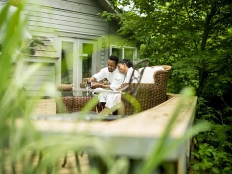 A couple toasts with wine glasses in white robes on a wicker couch outdoors.