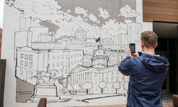 A young man takes a photo of a black and white line drawing mural of Columbia's skyline on his cellphone.