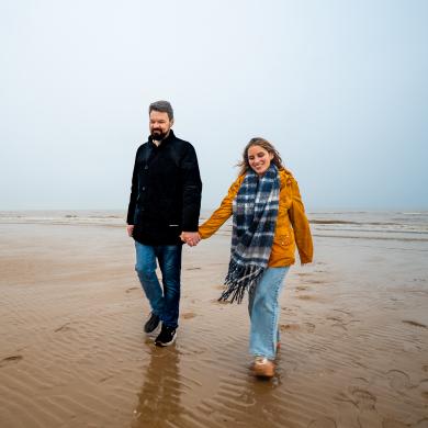 A couple wearing coats and scarves walk along a sandy beach