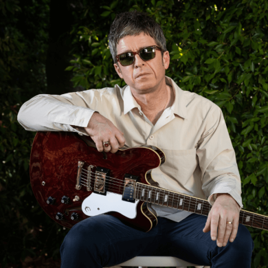 Noel Gallagher holding a guitar sitting in front of a hedge.