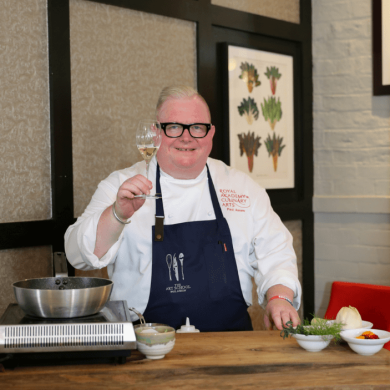 Chef Paul Askew stood at a workspace doing a cooking demonstration holding a glass of wine.