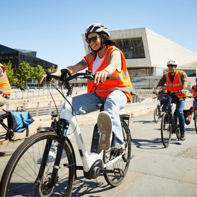 Four people cycling bikes along Pier Head with helmets on. The sun is shining and it's a beautiful day.