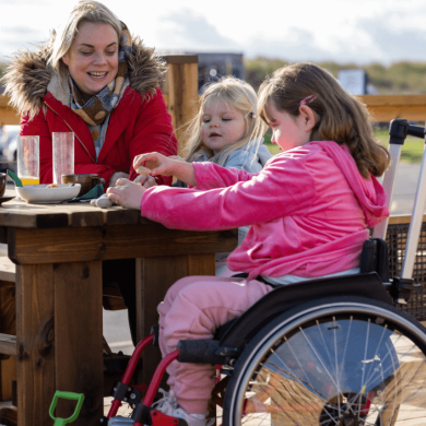 A family sitting by the seaside eating food, two children sit with their parents. One child is wearing a pink coat and is a wheelchair user.