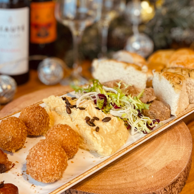 A collection of food on a long rectangle plate including pigs in blankets, falafel, humous and bread