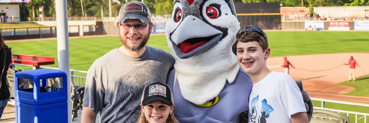 Family posing with a baseball team mascot at a stadium, smiling during a game-day event