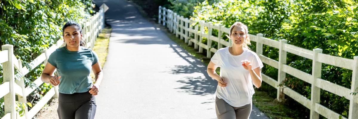 Two people jogging along the Cape Fear River Trail on a paved path lined with trees and white fencing.