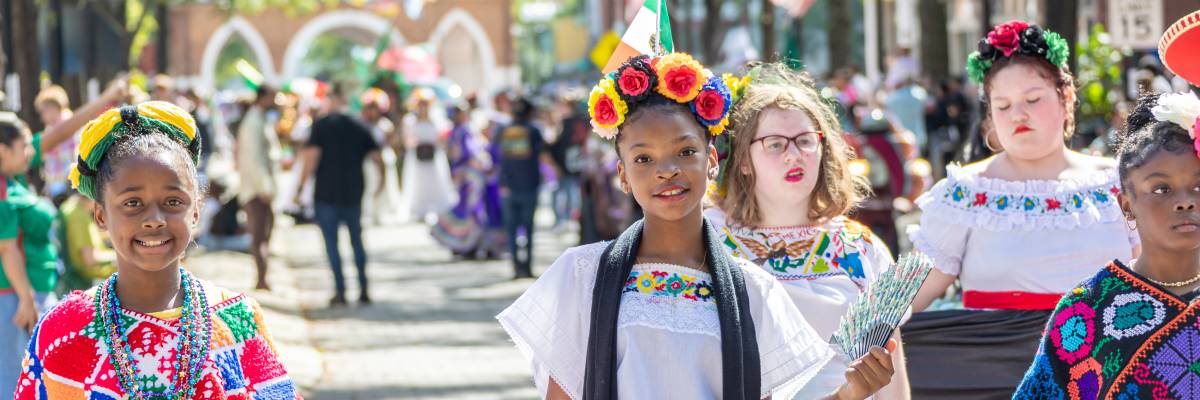 Group of children and teens in vibrant cultural dress participating in the International Folk Festival parade on Hay Street in Fayetteville, NC, with flags waving and crowds watching.