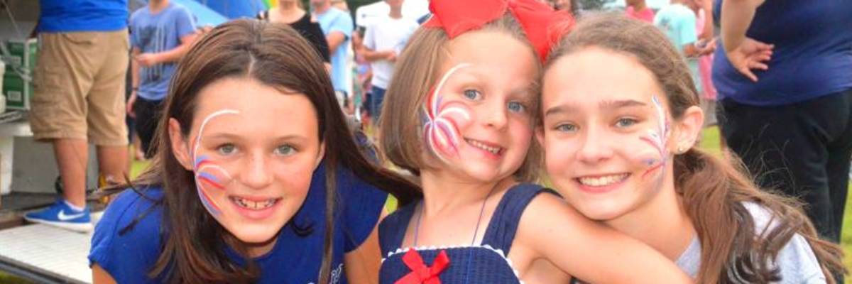 Three girls smiling together at the Stedman Fireworks Festival, with face paint and festival activities in the background.
