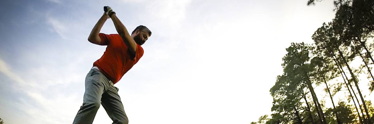 Golfer swinging a club on a grassy fairway surrounded by trees under an open sky