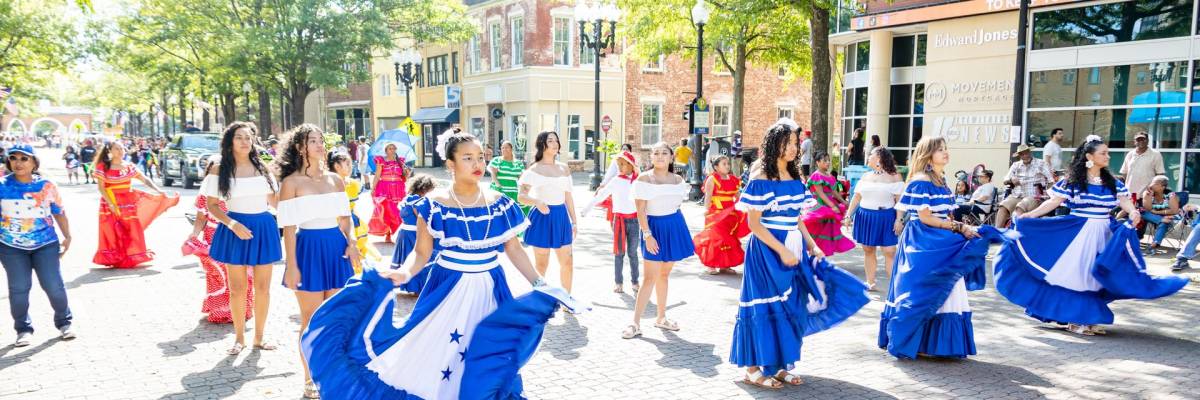 Dancers in blue and white dresses perform in downtown Fayetteville during a daytime cultural festival, with spectators lining the brick street.
