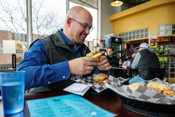 Man in a blue shirt and vest smiling at a sandwich at the Bashful Bison. A blue cup and menu are on the table. Others are dining in the background.