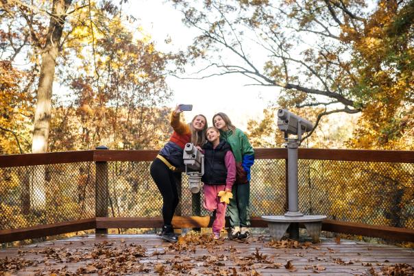 Family smiles and poses for a selfie outdoors during a fall day.
