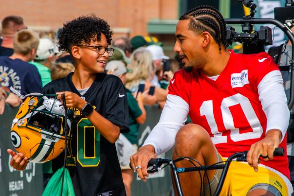 Jordan Love and a young boy at Packers Training Camp. Jordan is biking and the young boy is walking next to him carrying his helmet.