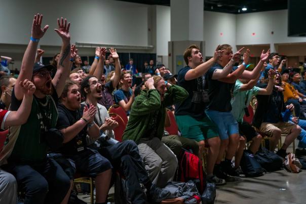 A crowd of attendees sitting in rows of chairs reacts with excitement, cheering, clapping, and raising their arms during an event inside a convention hall.