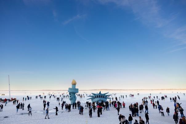 A wide shot of dozens of people walking on a frozen lake with a giant inflatable Lady Liberty in the center
