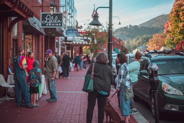 Poeple are gathered by vendor booths along brick-paved sidewalks with a vibrant-orange leaves on a nearby tree and a mountain ridge in the distance.