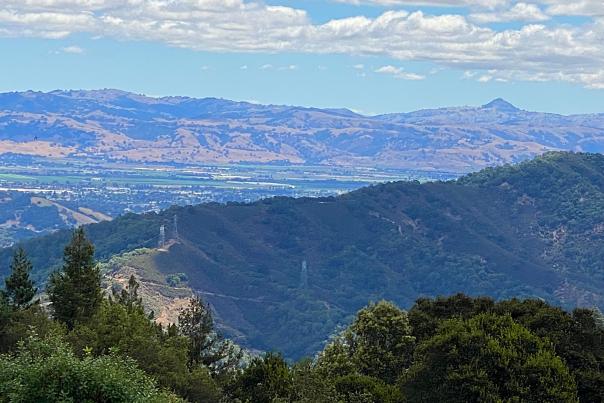 Elevated view of Gilroy looking eastward from Mt. Madonna Summit, showing redwood covered mountain range, the city resting in the valley below and the Diablo mountain range in the distance with blu sky and clouds above.
