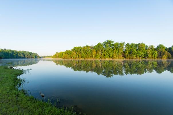 Image is of the Kincaid Lake in at sunset with the water in the foreground and trees and blue sky in the background.