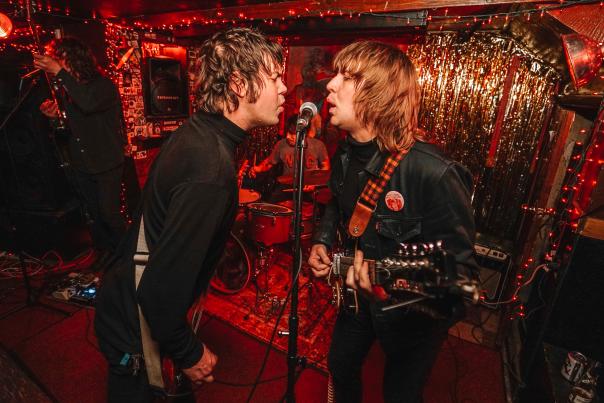 Two musicians  with guitars from the band The Whiffs lean into a microphone to sing.