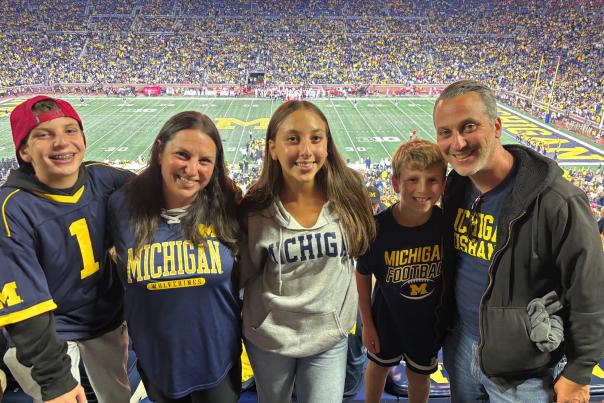 Family of five all wearing michigan sweatshirts and jerseys smile for the camera inside the Michigan Stadium, with the field and filled bleachers in the background.